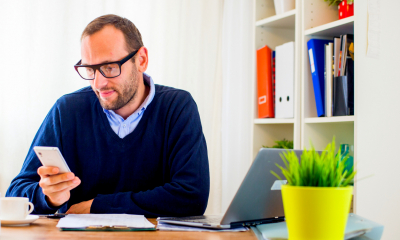Bearded man in a blue sweater wearing glasses looking at his phone sat at a desk
