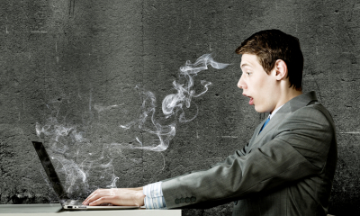 A man in a suit typing at a laptop that has smoke coming out of it