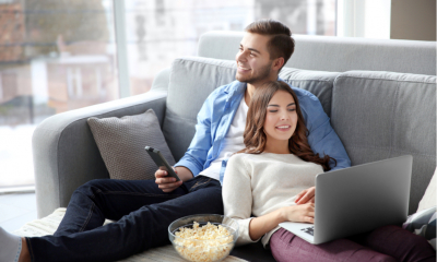 Couple watching TV and whilst woman multitasks using her laptop on a sofa at home